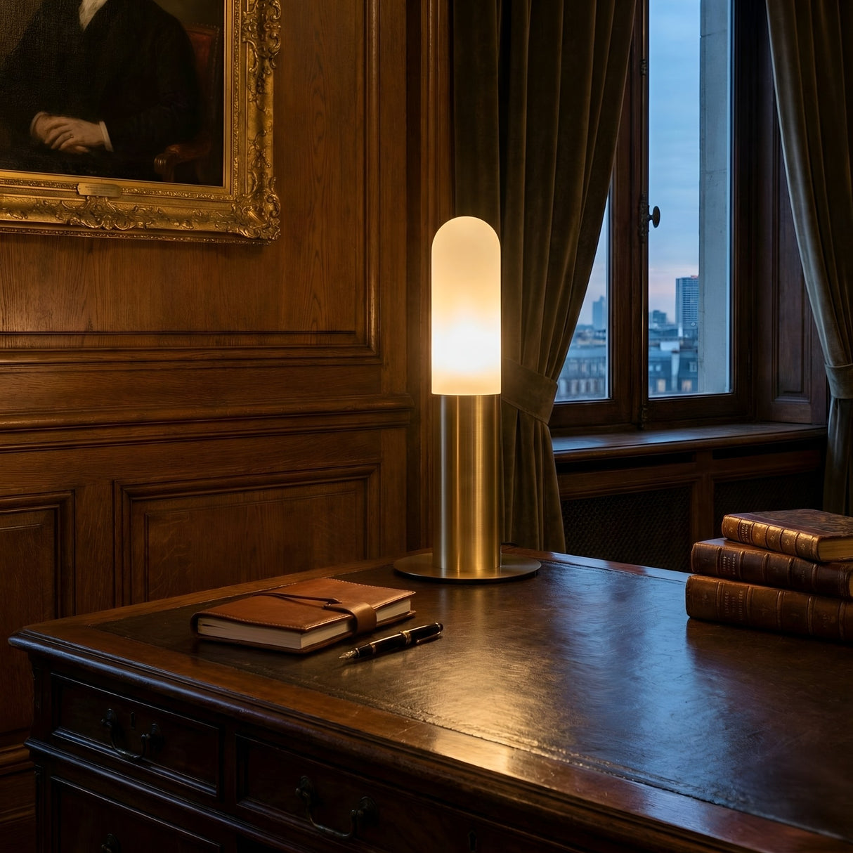 The Altessa 1 Light Small Vertical Alabaster Table Lamp in Soft White & Brass stands on a wooden desk beside stacked books, a closed notebook, and a pen, set in a wood-paneled room with curtains and a framed portrait.