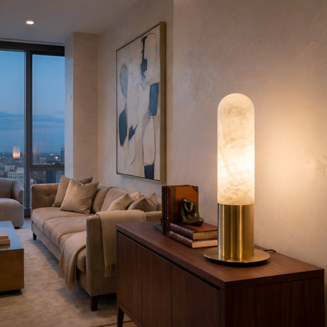 A modern living room at dusk has beige walls, a neutral-cushioned sofa, abstract art, a wooden console with books, and the Altessa 1 Light Small Vertical Alabaster Table Lamp in Soft White & Brass. Large windows showcase a cityscape view.