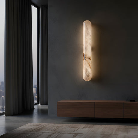 A modern interior features the Arvella LED Large Column Alabaster Wall Light in soft white, mounted on a dark wall beside a large window and low wooden cabinet, with city views visible through the glass.
