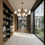 A modern hallway with tall bookshelves on the left, a floor-to-ceiling window on the right, beige tile flooring, and an elegant Ivy 18 Light Chandelier - Brass overhead, with a staircase leading to an upper floor in the background.