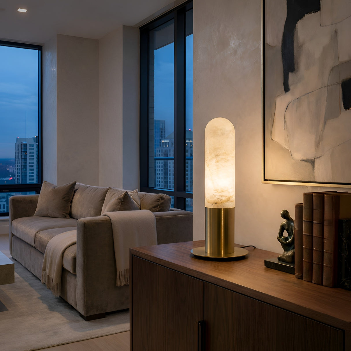 A modern living room at dusk with a beige sofa, city views from large windows, and a wooden sideboard displaying books, a sculpture, and the Altessa 1 Light Small Vertical Alabaster Table Lamp – Soft White & Brass softly glowing.