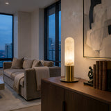 A modern living room at dusk with a beige sofa, city views from large windows, and a wooden sideboard displaying books, a sculpture, and the Altessa 1 Light Small Vertical Alabaster Table Lamp – Soft White & Brass softly glowing.