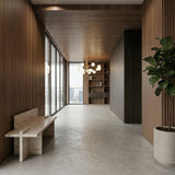 Modern hallway with wooden walls and ceiling, a minimalist bench, potted plant, and large windows bathing the space in light. An Ivy 18 Light Chandelier - Brass adds elegance above the built-in bookshelf in the background.