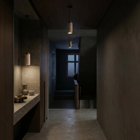 A dimly lit modern hallway with a concrete floor, dark walnut walls, and a window by the staircase features a Corano 1 Light Cylinder Travertine Single Pendant Light in Walnut Brown & Light Beige above a countertop with decorative objects.