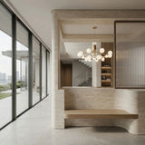 A modern interior features large floor-to-ceiling windows, a minimalist wooden bench, stone walls, and a staircase. The Ivy 18 Light Chandelier - Brass serves as a sculptural centerpiece with decorated shelves in the background.