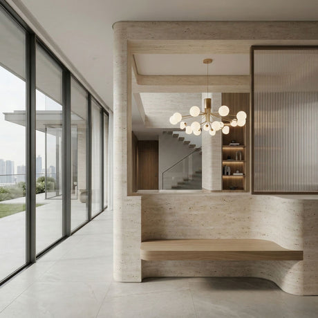 A modern interior features large floor-to-ceiling windows, a minimalist wooden bench, stone walls, and a staircase. The Ivy 18 Light Chandelier - Brass serves as a sculptural centerpiece with decorated shelves in the background.