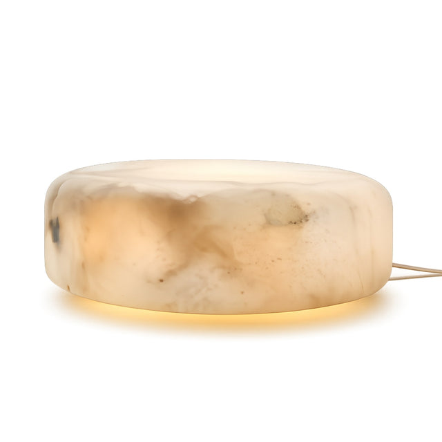 The Valenor 1 Light Disc Alabaster Table Lamp - Soft White features a round, low-profile design in natural Spanish alabaster with a marbled cream and beige finish, emitting a soft glow from below. Includes a power cord on the right side.