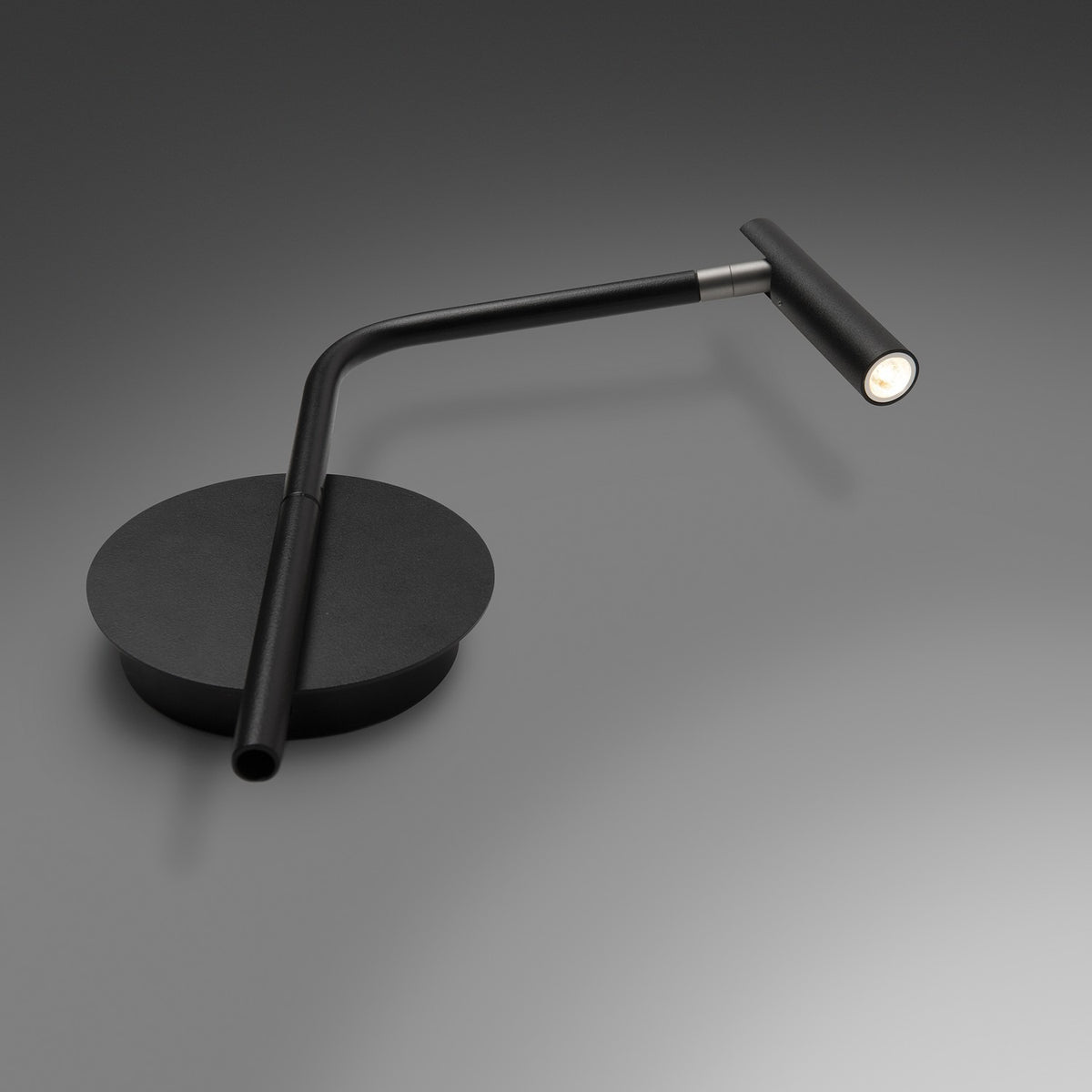 MiniSpot LED wall light in black iron with round wall plate, angled rod arm, and cylindrical spot head with grey collar joint, shown lit on a grey background.
