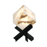 The Kalythra 1 Light Faceted Alabaster Table Lamp features a glowing, cube-shaped soft white alabaster shade with brown veining, supported by intersecting matte black metal legs in an X-shape for a modern geometric look.