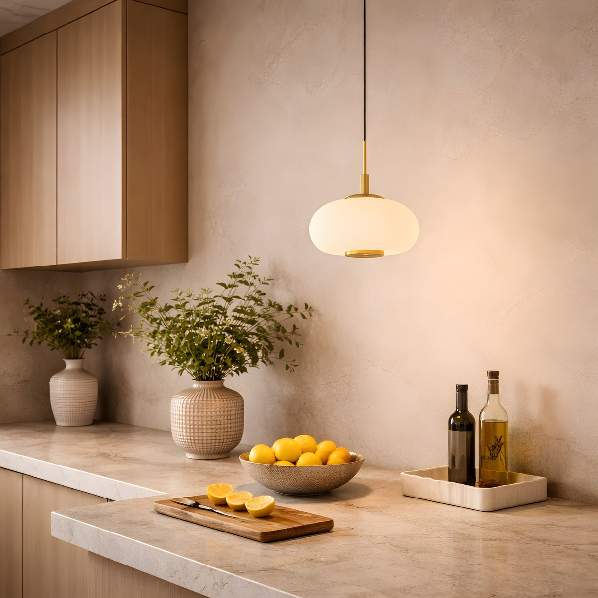 A modern kitchen countertop with a bowl of lemons, sliced lemons on a cutting board, bottles of oil, two potted plants, and the Aurevia LED Dome Alabaster Single Pendant Light – Brushed Brass against beige walls and wooden cabinets.