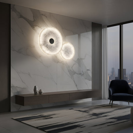 A modern living room with marble walls, a floating wooden console, decorative vases, and the Solmira LED Dual Disc Alabaster Wall Light in soft white & brushed brass. Floor-to-ceiling windows frame a cityscape at dusk. A dark armchair sits by a patterned rug.