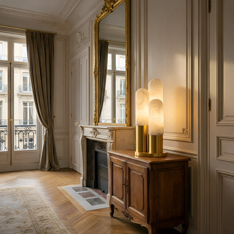Elegant living room with tall windows, a gold-detailed mirror above a marble fireplace, beige curtains, hardwood floors, and a wooden sideboard topped with the Altessa 3 Light Vertical Alabaster Table Lamp in Soft White & Brass.