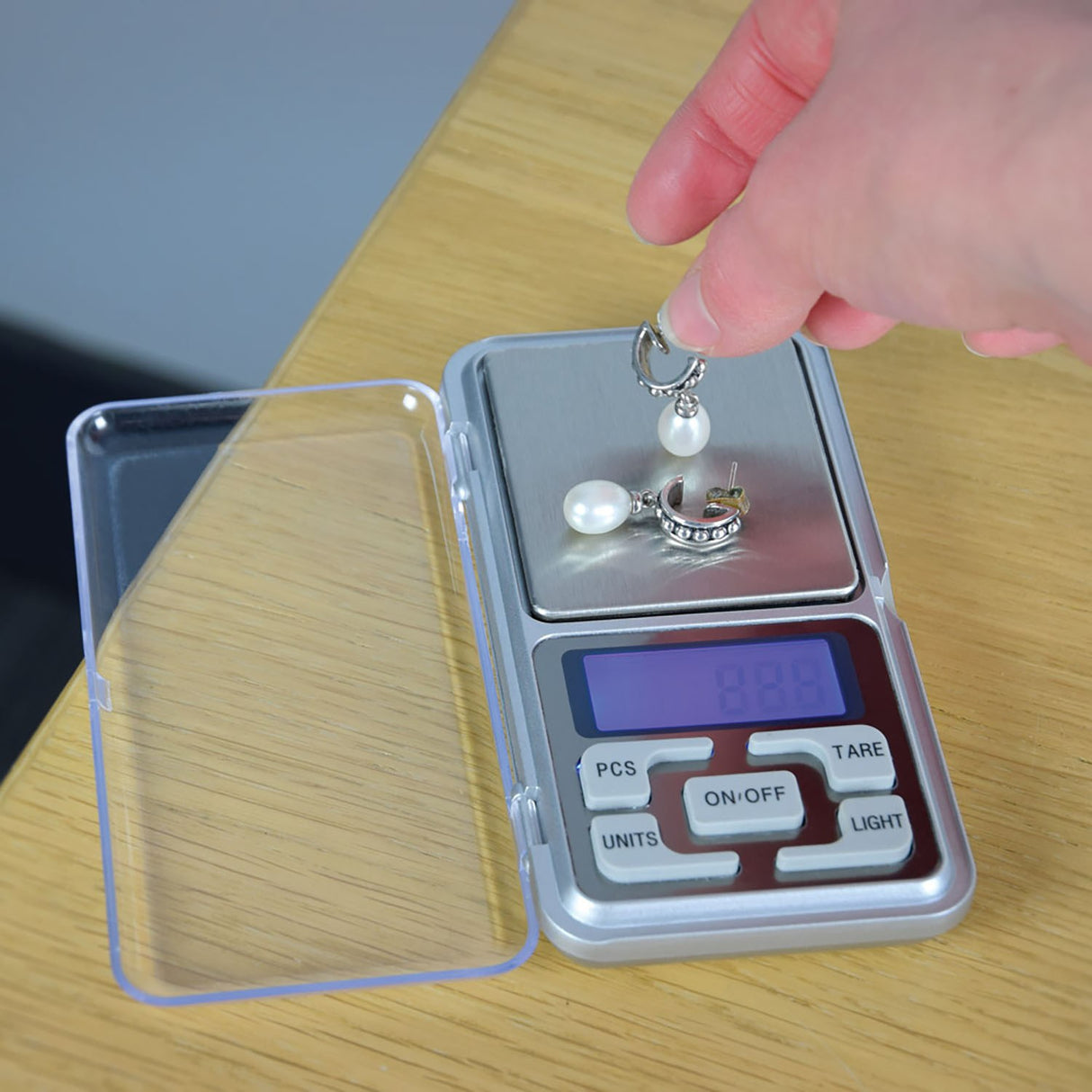 AVSL Mercury digital pocket scale open on a wooden surface with clear flip cover, LCD blue backlight, and hand placing pearl earrings on the platform.