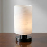 The Cylorin LED Cylindrical Alabaster Table Lamp in soft white and black sits on a wooden surface, softly glowing against a plain light gray background with its modern cylindrical shade and round black base.
