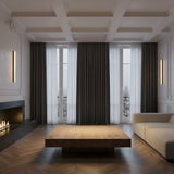 A modern living room features a large wooden coffee table, beige sofa, sleek fireplace, tall windows with sheer and dark curtains, herringbone wood floors, and the Cornova LED Medium Slim Linear Alabaster Wall Light in soft white & matte black for a minimal look.