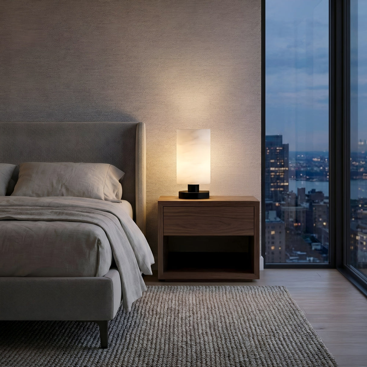 A modern bedroom with a grey upholstered bed, beige bedding, a wooden nightstand featuring the Cylorin LED Cylindrical Alabaster Table Lamp in Soft White & Black, a textured rug, and large windows overlooking the city skyline at dusk.