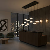 Niori Aurevia 14 Light LED linear chandelier in matt black and brass with staggered alabaster domes above a wood reception desk by a city-view window.