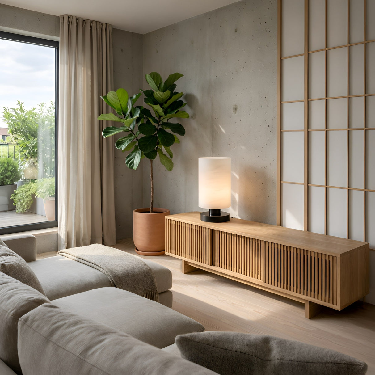 A modern living room with a beige sofa, wooden media console, potted plant, Cylorin LED Cylindrical Alabaster Table Lamp in Soft White & Black, floor-to-ceiling window with curtains, and sliding shoji screen embraces minimalist style.