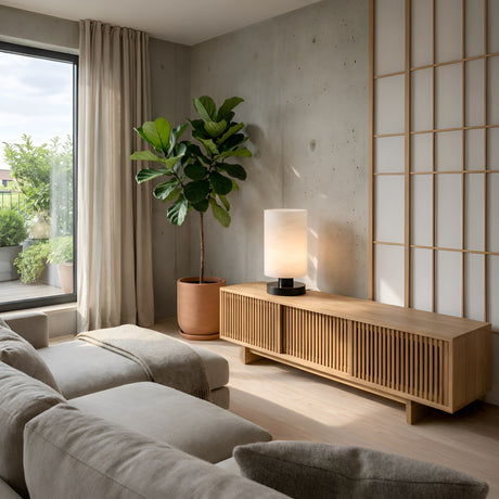 A modern living room with a beige sofa, wooden media console, potted plant, Cylorin LED Cylindrical Alabaster Table Lamp in Soft White & Black, floor-to-ceiling window with curtains, and sliding shoji screen embraces minimalist style.