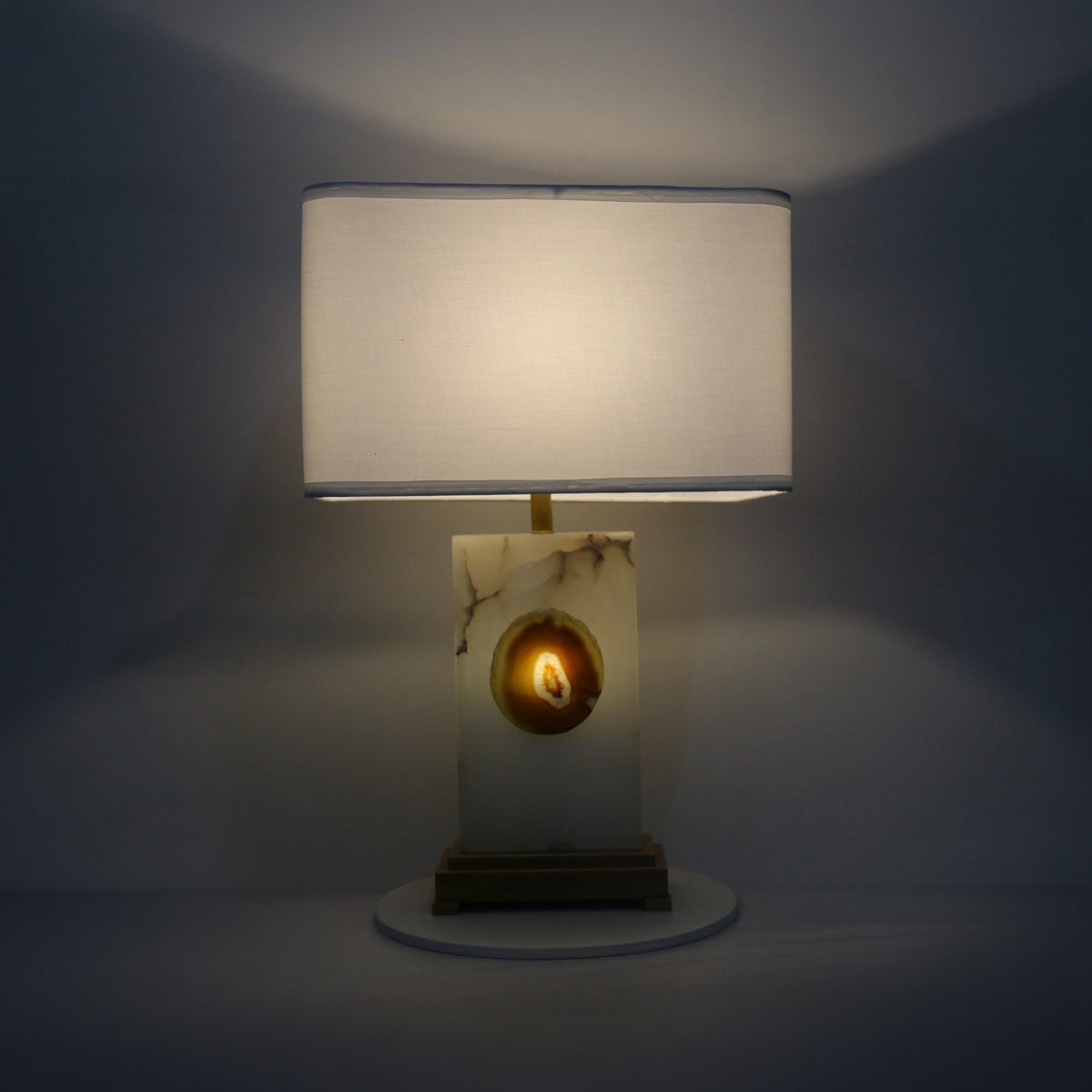 Selune alabaster table lamp lit at night, showing rectangular fabric shade, brushed brass stem and plinth, and glowing agate slice inset.