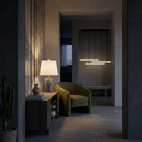A cozy, modern room showcases a green armchair next to a wooden table topped with the Marivon 1 Light Marble Cylinder Table Lamp in Brass & Grey. Minimal decor includes a cactus, and pendant lights softly illuminate the hallway.