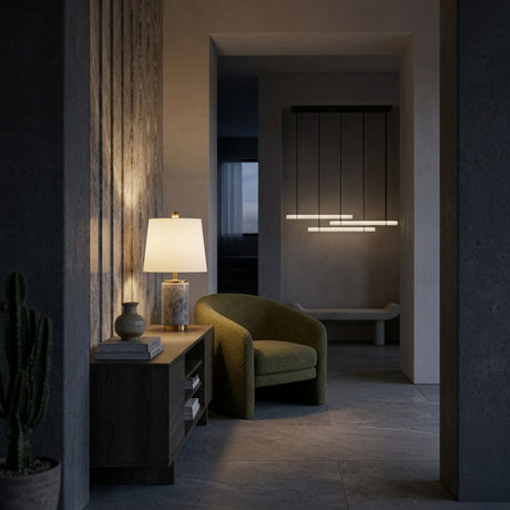 A cozy, modern room showcases a green armchair next to a wooden table topped with the Marivon 1 Light Marble Cylinder Table Lamp in Brass & Grey. Minimal decor includes a cactus, and pendant lights softly illuminate the hallway.