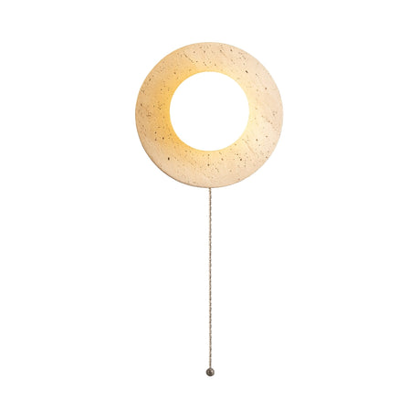 The Solenne 1 Light Travertine Wall Light With Pull Switch in light beige features a round, textured design and emits a warm glow from its circular center. A small pull switch hangs below, making it perfect for ambient lighting against any wall.