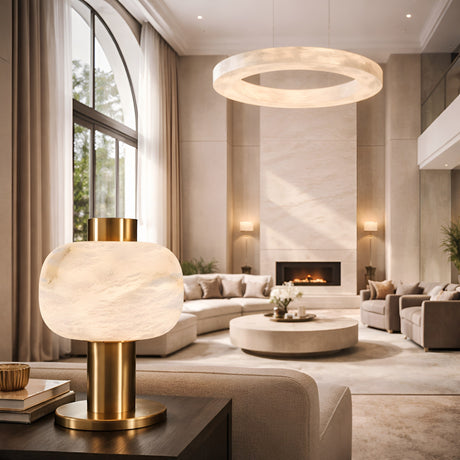 A modern living room with tall arched windows, neutral furniture, a round coffee table, fireplace, and elegant lighting featuring the Marveta 1 Light Small Rounded Alabaster Table Lamp in soft white & brass beside a circular ceiling light.