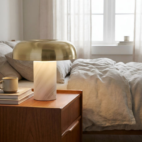 A modern bedroom with an unmade bed in light beige bedding, a wooden nightstand holding books, a ceramic mug, and the Arven LED Mushroom Form Marble Table Lamp in soft white & gold beside a window with sheer curtains.