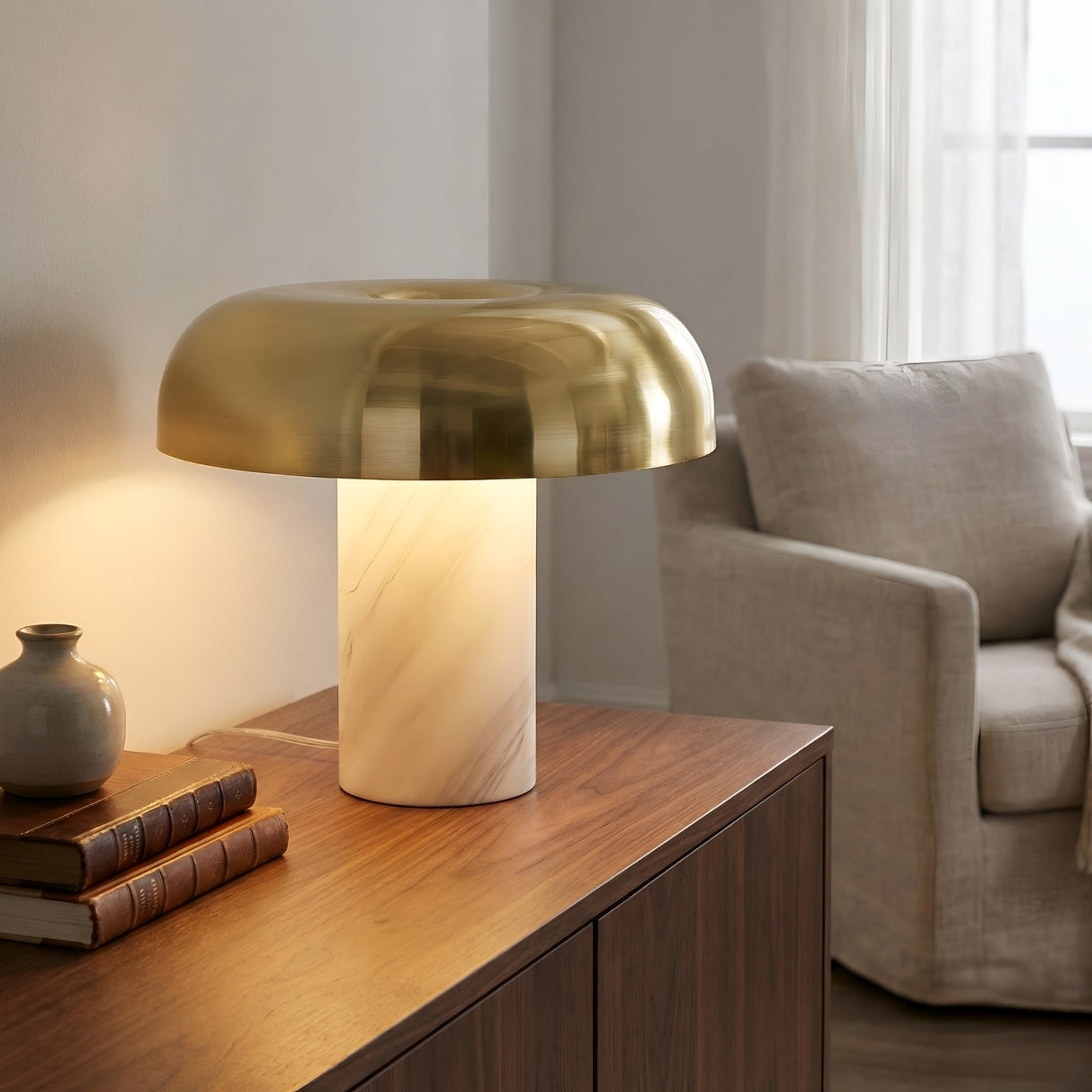The Arven LED Mushroom Form Marble Table Lamp in Soft White & Gold rests on a wooden sideboard next to books and a gray vase, with a beige armchair and softly lit window in the background.