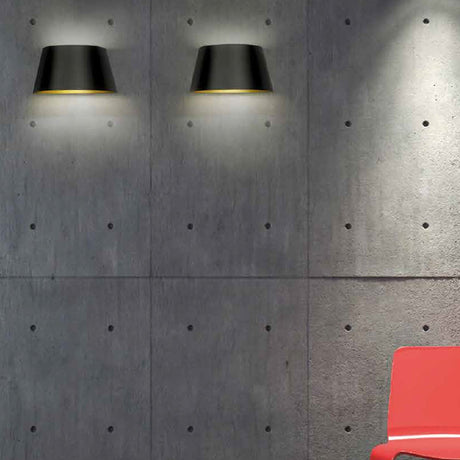 Fennorne Wall Light in black with gold interior, two tapered iron fixtures mounted on a concrete wall, glowing gold at the rim with light cast downward, red chair below.