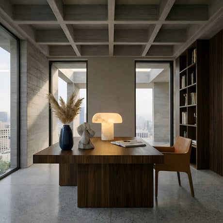 A modern office features a wooden desk, leather chair, sculptural decor, and the Calvessa LED Solid Alabaster Mushroom Table Lamp in Soft White glowing on the desk; floor-to-ceiling windows reveal a city view, while book-lined shelves line one wall.