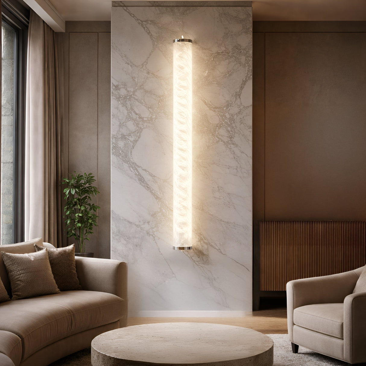 A modern living room with neutral tones, a curved beige sofa, round coffee table, armchair, and a Corvane LED Medium Slim Cylindrical Alabaster Wall Light in soft white on a marble accent wall, plus a plant and large window in the background.