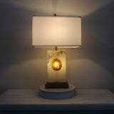 Selune table lamp on a bedside surface with alabaster base, glowing agate slice centre, brushed brass stem, stepped plinth, and white fabric shade.