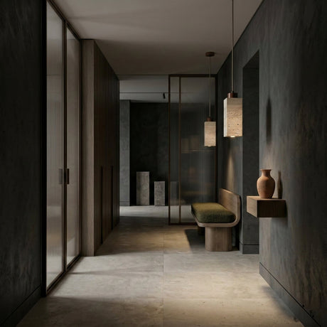 A minimalist hallway with dark walls features a wooden bench, a green cushion, and the Zerano 1 Light Rectangular Travertine Single Pendant Light in walnut brown & light beige. A wall shelf with a ceramic vase, glass partitions, and stone flooring complete the serene look.