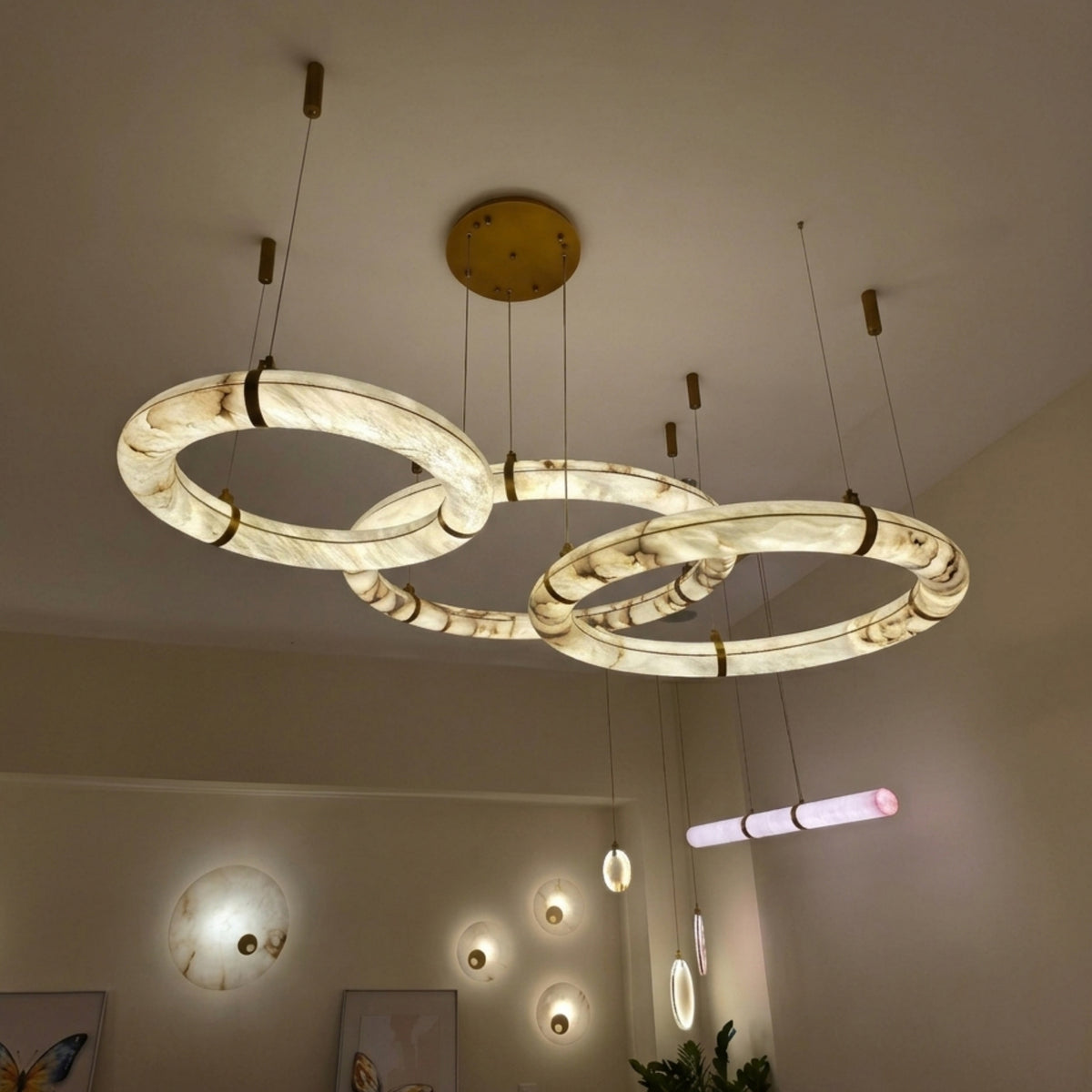 Aureon LED Triple Ring Alabaster Chandelier in brushed brass, three staggered glowing rings suspended from a round ceiling canopy in a warm-toned living space.