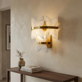 The Petraia LED Floral Alabaster Wall Light in brushed brass and soft white casts a warm glow above a wooden console with a vase of dried branches and books, set against a beige textured wall.