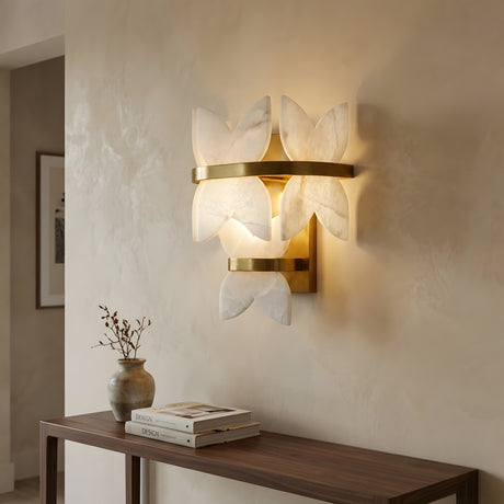 The Petraia LED Floral Alabaster Wall Light in brushed brass and soft white casts a warm glow above a wooden console with a vase of dried branches and books, set against a beige textured wall.