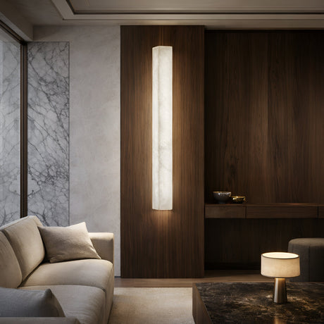 A modern living room with a beige sofa, marble-topped table, and Valmera LED Large Slim Vertical Alabaster Wall Light (Soft White) on wood paneling; a table lamp and marble-patterned wall complete the minimalist elegance of the space.