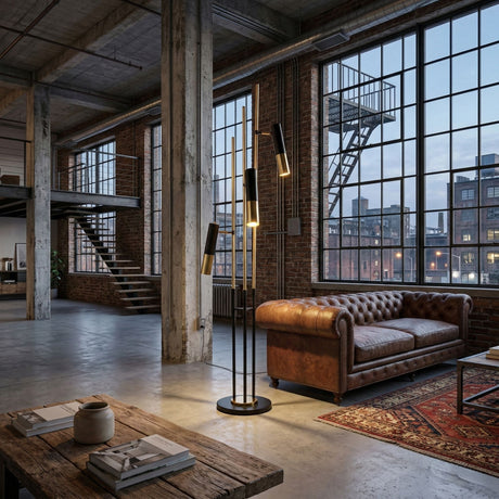 A modern industrial loft features large factory windows, a brown leather sofa, a patterned rug, and the Leonard 3 LED Floor Lamp - Black & Gold for energy-efficient lighting. Exposed brick walls and concrete floors complete the urban decor.