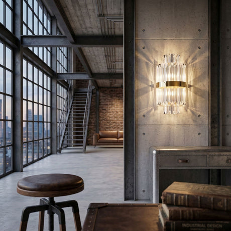 A modern industrial loft with large grid windows, exposed pipes, a concrete wall featuring the Liberty 2 Light Crystal Glass Wall Light in gold, a brown leather sofa, metal cabinet, wooden stool, and stacked books on a table.