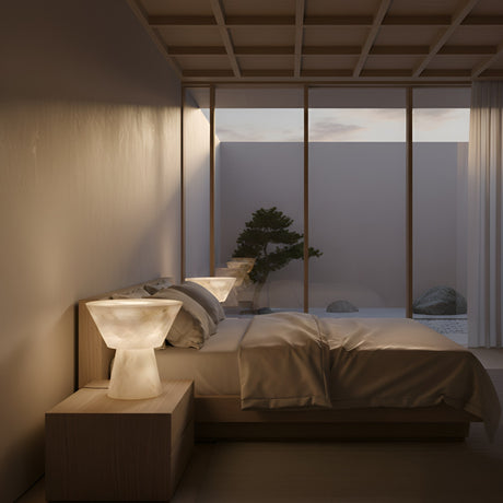 A minimalist bedroom features a low wooden bed with white bedding and two Calyrix LED Small Flared Alabaster Table Lamps in soft white. A large window overlooks a rocky garden with a bonsai, filling the space with gentle natural light.