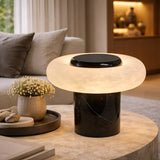 The Orvixa LED Rounded Alabaster Disc Table Lamp in Soft White & Black sits on a round coffee table beside a small vase of white flowers and smooth stones, illuminating a cozy, softly lit living room.