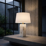 The Kavryn 1 Light Raw Cut Alabaster Table Lamp in Brass & Soft White, featuring a rectangular textured stone base and fabric shade, sits on a concrete surface in a minimalist room with large windows and concrete walls.