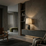 A modern, minimalist living room with dark textured walls, a floating console table, wooden chairs, built-in shelves with vases, and a green bench next door. Warm lighting and the Valmera 1 Light Cylinder Marble Table Lamp - Brass & Black create a cozy vibe.