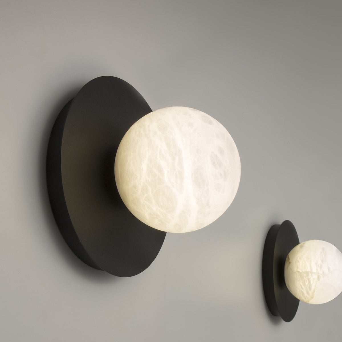 Halvessa LED Alabaster Dimmable Wall Light in Black, with a round globe set off-centre on a black iron disc, shown lit against a grey wall alongside a second matching light.