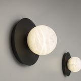 Halvessa LED Alabaster Dimmable Wall Light in Black, with a round globe set off-centre on a black iron disc, shown lit against a grey wall alongside a second matching light.