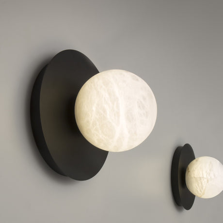 Halvessa LED Alabaster Dimmable Wall Light in Black, with a round globe set off-centre on a black iron disc, shown lit against a grey wall alongside a second matching light.