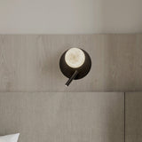 Halvessa LED Alabaster Wall Light with Reading Light in black, round iron plate with glowing alabaster globe and slim angled reading arm, shown above a bed.