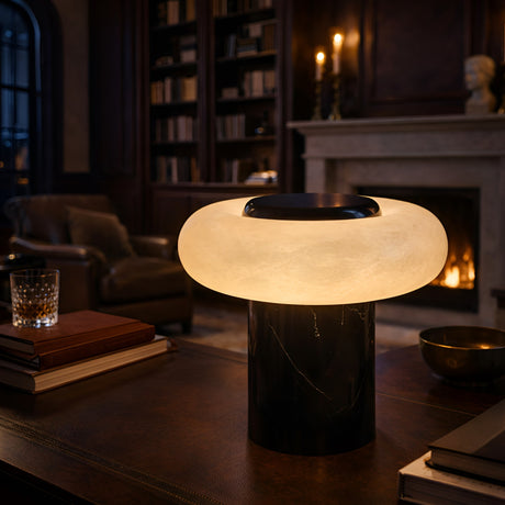 The Orvixa LED Rounded Alabaster Disc Table Lamp in Soft White & Black glows atop a wooden table in a cozy library with dark wood shelves, armchairs, a fireplace, and stacks of books.